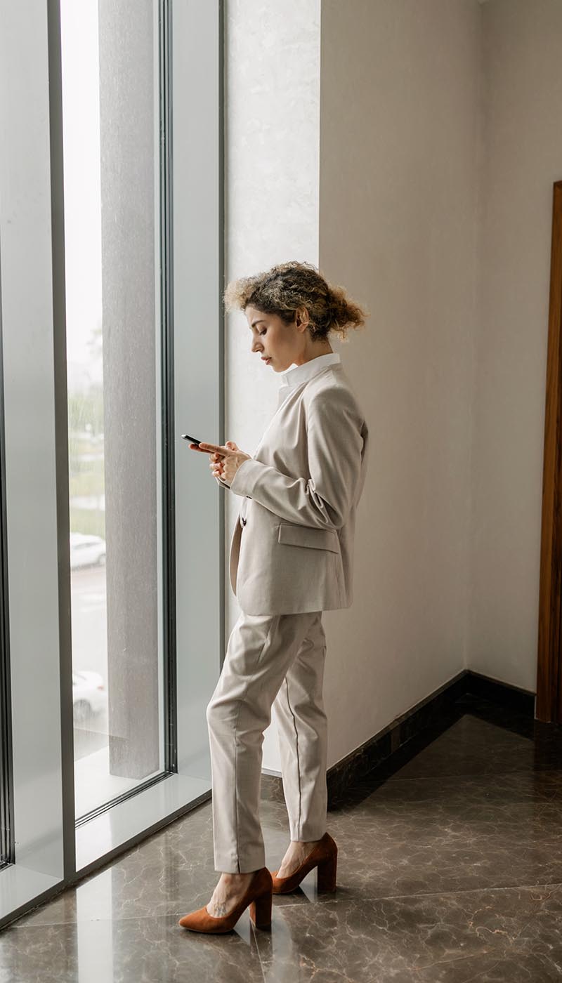 woman with curly hair wearing suit standing by office window looking at broker opinion of value report on phone woman with curly hair wearing suit standing by office window looking at broker opinion of value report on phone